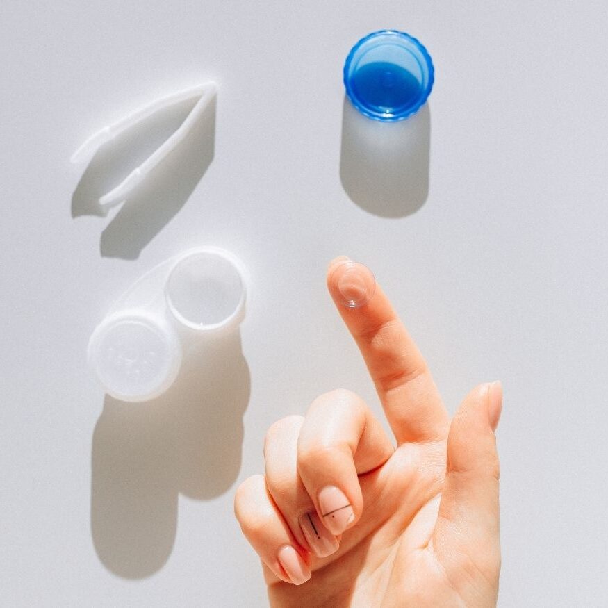Minimalist flat lay of contact lens care essentials including a white case, solution cap, tweezers, and a finger ready for lens application.