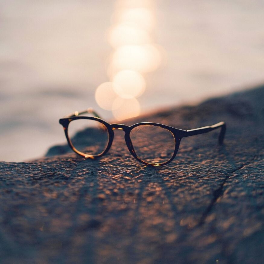 Artistic close-up of a pair of black-rimmed eyeglasses resting on a textured rock at sunset with golden bokeh light reflecting off the water.