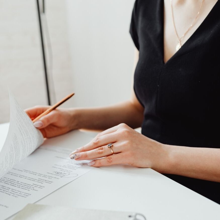 Close-up of a professional woman in a black blouse signing a document on a white desk in a bright, minimalist office setting.